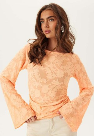 Donna con capelli castani ondulati che indossa una maglia pescata a maniche lunghe con trama floreale, polsini svasati e grandi orecchini d'argento, guarda di lato.