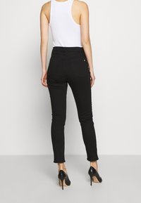 Mujer vistiendo jeans negros ajustados, top sin mangas blanco y tacones altos negros, de pie con la espalda hacia la cámara contra un fondo blanco.