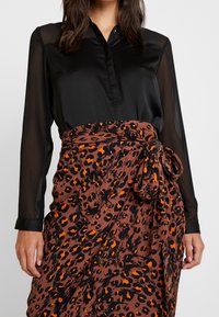 Femme portant un chemisier noir transparent à manches longues rentré dans une jupe portefeuille imprimée léopard marron et orange nouée à la taille.