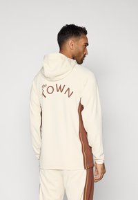 Capuchontrui in crème met bruine zijpanelen en de tekst "The TOWN" op de rug. Glad van textuur met lange mouwen en een relaxte pasvorm.