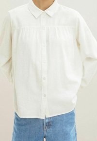 Camicia bianca con bottoni realizzata in tessuto leggero, caratterizzata da un colletto, pieghe frontali e una leggera texture, abbinata a jeans blu.