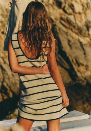 Woman with wavy hair wearing a sleeveless black-and-white striped dress standing outdoors with rocky background facing away.