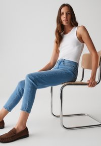 Jean en denim bleu clair avec une taille haute, coupe slim et détails de décoloration subtils, associés à un débardeur blanc à côtes et des mocassins marron.