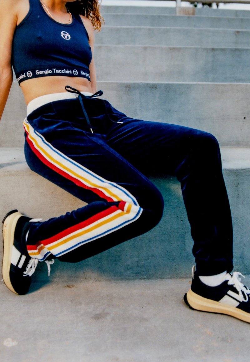 Marineblaue Velours-Trackpants mit weißen, roten, gelben und blauen Streifen an den Seiten, kombiniert mit schwarz-weißen Sneakern.
