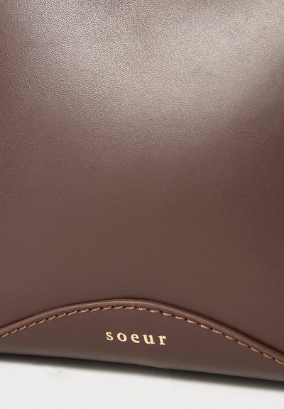 Ruskea nahkalaukku, jossa on sileä pinta, kaareva sauma ja kultaisella kohokuvioidulla "soeur"-brändin nimellä varustettu.