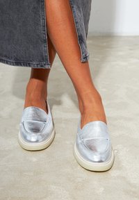 Metallisch-silberne Slippers mit abgerundetem Zehenbereich und flacher Sohle. Glatte Textur mit einem dezenten Glanz, kombiniert mit einer hellen Außensohle.