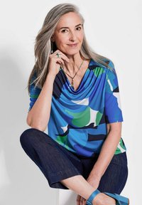Bunt gemusterte Bluse mit einem drapierten Ausschnitt, mit blauen, grünen und weißen abstrakten Mustern, kombiniert mit dunkelblauen, knöchellangen Hosen und blauen Sandalen.