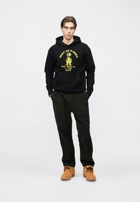 Čierna mikina s kapucňou so žltým grafickým textom, kombinovaná s tmavozelenými manšestrovými nohavicami a hnedými pracovnými topánkami. Model stojí na svetlom pozadí.