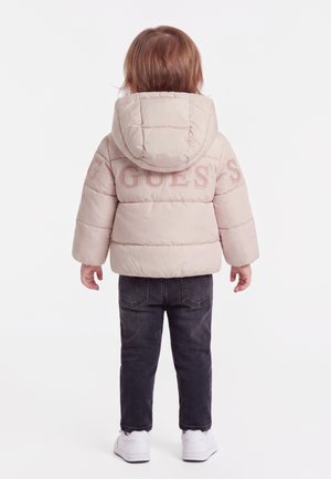 Enfant aux cheveux roux portant une doudoune rose clair avec capuche et un jean, debout de dos sur fond blanc.