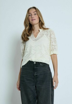 Lace blouse in pale yellow with a v-neck and puffed sleeves, paired with high-waisted dark denim jeans. The fabric features floral patterns.