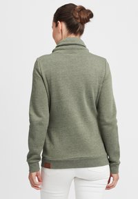 Olivenfarget genser med høy rullet krage, lange ermer og strukturert stoff. Har en brun lærlogo-patch nederst.
