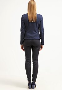 Blazer ajusté bleu marine à manches longues, tissu texturé. Associé à un pantalon noir ajusté et des chaussures à talons blocs bleu marine. Vue de dos.