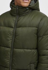 Grøn pufferjakke med hætte, der har en frontlynlås og quiltet design. Lavet af skinnende, glat materiale med en afslappet pasform.