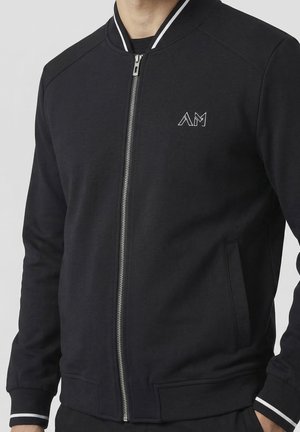 Homme portant une veste bomber noire zippée avec des bordures blanches au col et aux poignets, et un petit logo blanc "AM" sur la poitrine gauche.