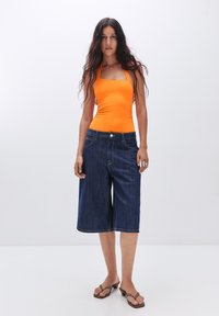 Oranje nauwsluitend tanktop gecombineerd met donkerblauwe denim culottes, voorzien van een knoopsluiting en een ruwe zoom. Het model draagt bruine flip-flops.