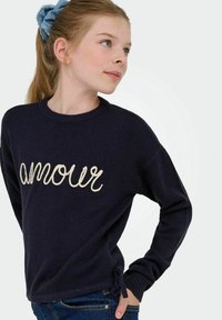 Mala deklica z svetlo rjavimi lasmi, vezanimi z modrim scrunchijem, oblečena v mornarsko modro pulover s besedo "amour" in modre kavbojke, ki gleda vstran.