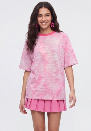 Ung kvinde med langt mørkt hår smiler og har pink stjerneøreringe, en lyserød tie-dye oversized t-shirt og et plisseret lyserødt skørt på.
