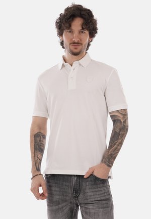 Uomo con capelli ricci che indossa una polo bianca e jeans grigi, con tatuaggi dettagliati su entrambi gli avambracci, una mano in tasca.