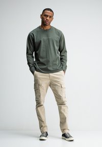 Man staat met zijn handen in zijn zakken, draagt een groene longsleeve shirt met tekst, beige cargobroek en zwarte hoge sneakers voor een witte achtergrond.