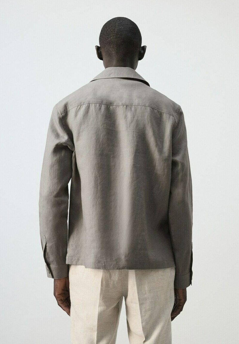 Homme debout, dos à la caméra, portant une chemise grise à manches longues et un pantalon beige clair, sur un fond clair uni.