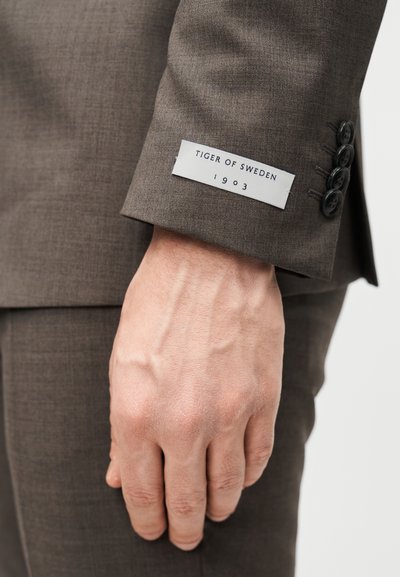 Το μανίκι του καφέ κοστουμιού έχει υφή υφάσματος, τέσσερα μαύρα κουμπιά και μια λευκή ετικέτα με την επιγραφή "TIGER OF SWEDEN 1903" σε ναυτικό μπλε χρώμα.