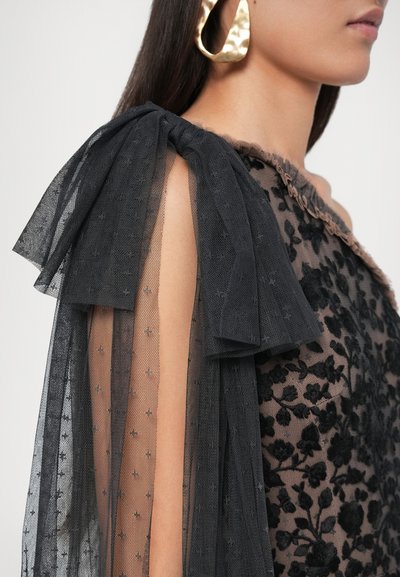 Umărul unei femei acoperit cu o manșetă din dantelă neagră florală, transparentă, legată într-o fundă mare, iar o cercelă texturată din aur vizibilă.