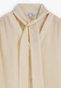 Blusa de un beige claro hecha de un tejido texturizado que presenta un cuello atado, un cierre frontal con botones y un diseño suave y fluido. La etiqueta dice "SCALPERS", tamaño S.