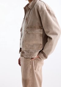 Beige denimjakke med afslappet pasform, store forlommer og synlige syninger. Båret over en hvid top og matchende beige bukser.