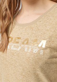 Beige tričko s krátkým rukávem a kulatým výstřihem. Obsahuje zlatý a bílý potištěný text a texturovaný design látky.