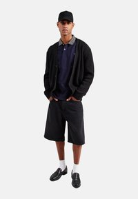 Cardigan de punto negro sobre un polo azul marino con cuello estampado, combinado con pantalones cortos negros, calcetines blancos y zapatillas de tela negras.