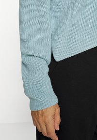 Hellblauer Strickpullover mit strukturierter Oberfläche und gerippten Bündchen. Schwarzer Stoff darunter sichtbar, mit glatter Oberfläche.