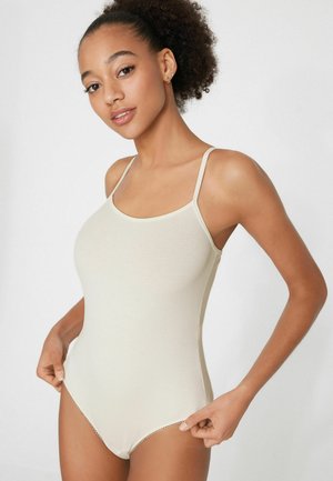 Młoda kobieta o kręconych włosach w kremowym prążkowanym body na cienkich ramiączkach, poprawiająca jego boki na jednolitym tle.
