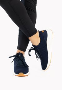 Scarpe da ginnastica blu navy con tomaia in maglia, suola intermedia bianca e suola in gomma. Sono visibili i lacci e una piccola etichetta di accento marrone.