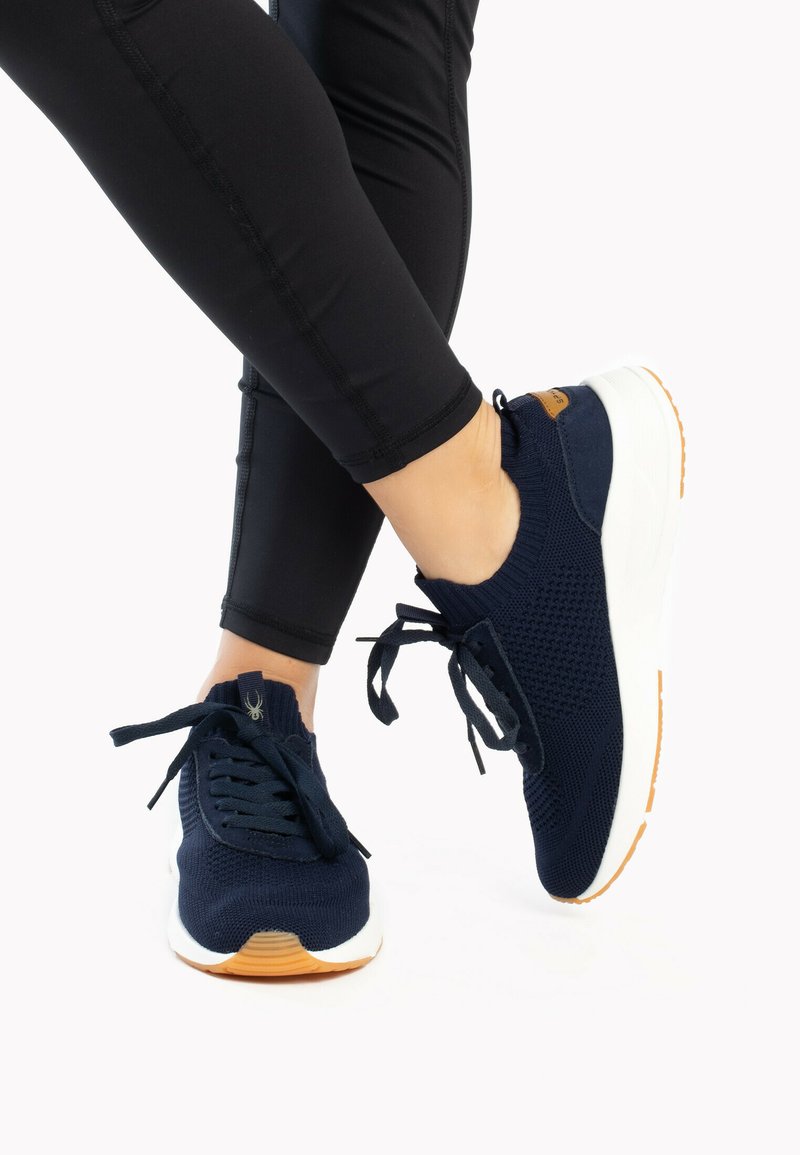 Scarpe da ginnastica blu navy con tomaia in maglia, suola intermedia bianca e suola in gomma. Sono visibili i lacci e una piccola etichetta di accento marrone.