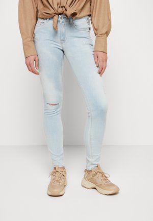 Persoon met lichtblauwe gescheurde skinny jeans, een beige geknoopte blouse en beige chunky sneakers, staand tegen een witte achtergrond.