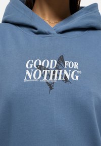 Blue hoodie with a soft texture, featuring a white print saying "GOOD FOR NOTHING" and a black butterfly design beneath.