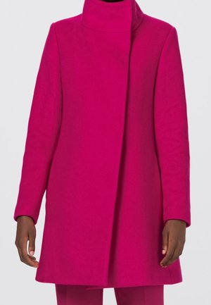 Manteau rose vif à manches longues avec col montant, fermeture avant dissimulée, porté par une personne avec les mains visibles sur les côtés.