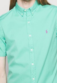 Košile na knoflíky s krátkým rukávem v mátově zelené barvě z bavlny, s růžovým vyšitým logem na levé straně hrudi a bílými knoflíky na přední straně.