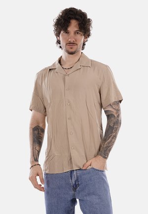 Hombre con cabello rizado que lleva una camisa beige de manga corta abotonada y pantalones vaqueros azules, mostrando tatuajes en ambos antebrazos, posando con una mano en el bolsillo.