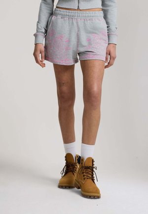 Osoba nosząca szare spodenki z różowymi kwiatowymi wzorami, białe skarpetki, beżowe sznurowane buty oraz szarą bluzę zapinaną na zamek, stojąca na neutralnym tle.