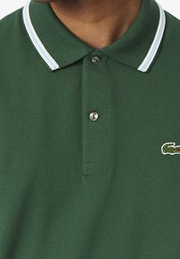 Zielona koszulka polo z teksturowanej tkaniny, z dwukolorowym kołnierzykiem w jasnoniebieskie paski oraz z markowym logo na piersi.