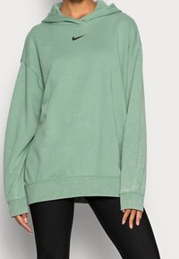 Person som bär en överdimensionerad ljusgrön Nike-hoodie och svarta leggings, stående mot en enfärgad grå bakgrund.