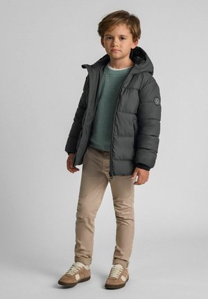 Pufferjakke i mørkegrå med hætte, med lynlåslukning og syning detaljer. Sammen med en grøn struktureret sweater og beige bukser.