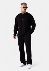 Schwarzes langärmliges Shirt mit Rundhalsausschnitt und kleinem weißen Logo, kombiniert mit schwarzen Hosen und weißen Turnschuhen. Glatte Stoffstruktur.