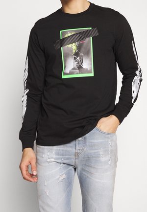 Mann trägt schwarzes Langarmshirt mit neon-grün gerahmtem Statuengrafik und weißen flammenähnlichen Streifen an den Ärmeln, kombiniert mit hellen, zerrissenen Jeans.