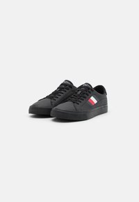 Zapatillas negras hechas de un material sintético liso, con una suela de goma texturizada y tres franjas de colores (rojo, blanco, azul) en el lateral.
