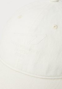 Casquette en coton de couleur blanc cassé avec un tissu texturé, logo brodé dans une couleur assortie, et accents cousus sur la visière et la couronne.