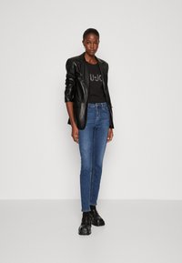 Blazer de cuero negro sobre una camiseta negra con logotipo de strass, combinado con jeans ajustados azules y zapatos gruesos negros. Fondo minimalista.