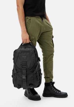 Persona con pantalones cargo verdes y botas negras sostiene una mochila negra por la asa superior contra un fondo liso.