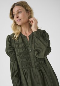 Abito verde, con bottoni e maniche a sbuffo, con dettagli smock sul corpetto e un tessuto morbido e testurizzato. Design elegante e fluido.